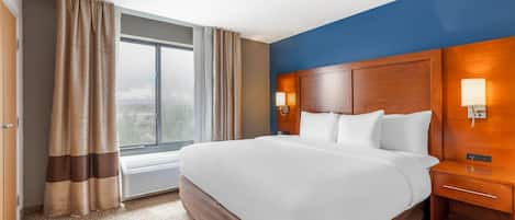 Habitación de hotel con una cama grande, un cabecero de madera, dos mesitas de noche con lámparas, una ventana con cortinas y vistas a la ciudad.