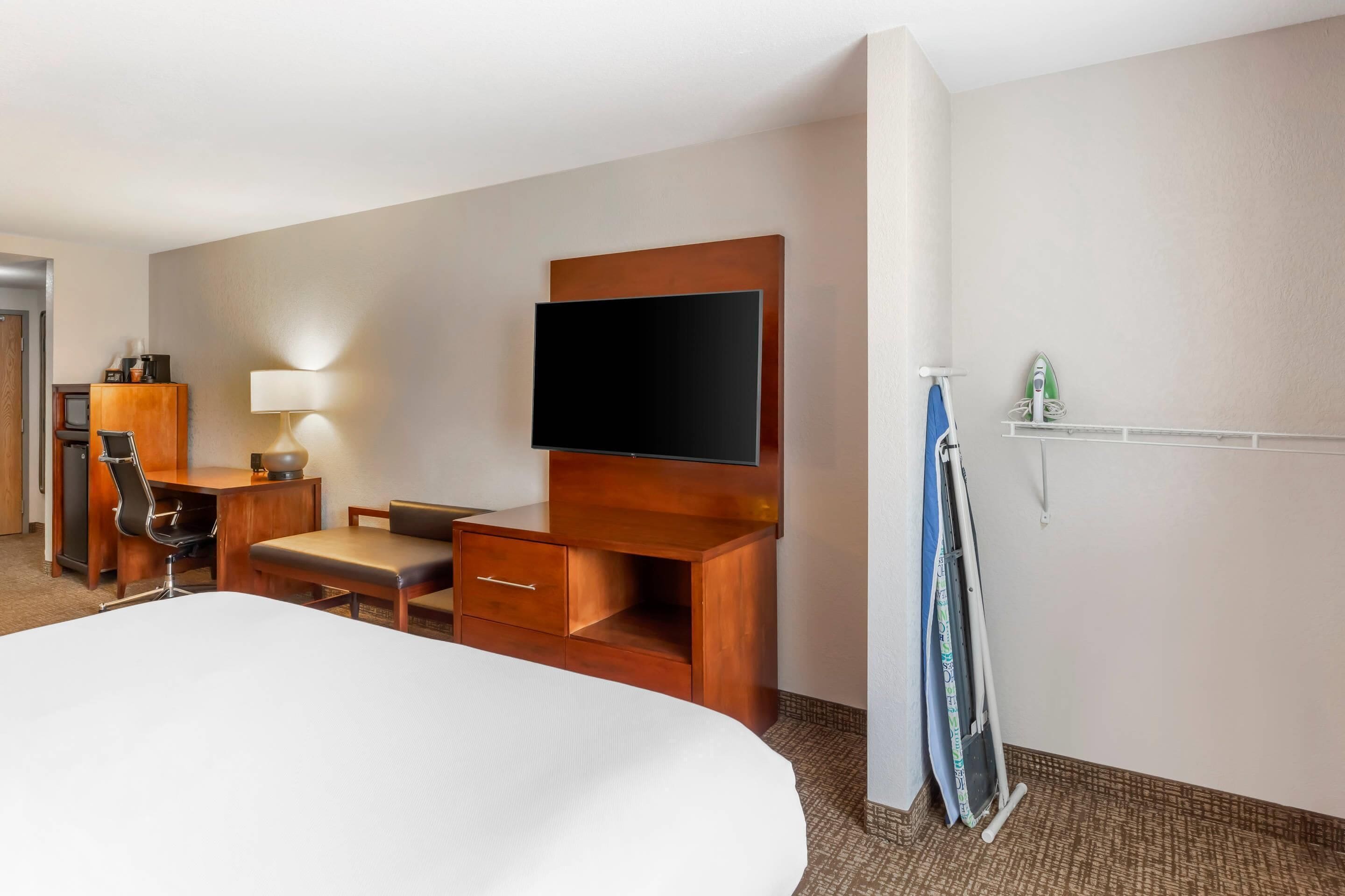 A hotel room with a large bed, a desk with a chair, a flat-screen TV, a lamp, and a ironing board.