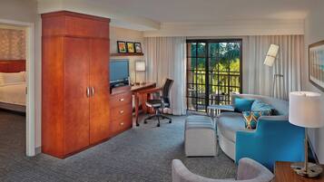 A hotel room with a bed, a desk with a computer, a sofa, a chair, and a large window with a view of greenery.