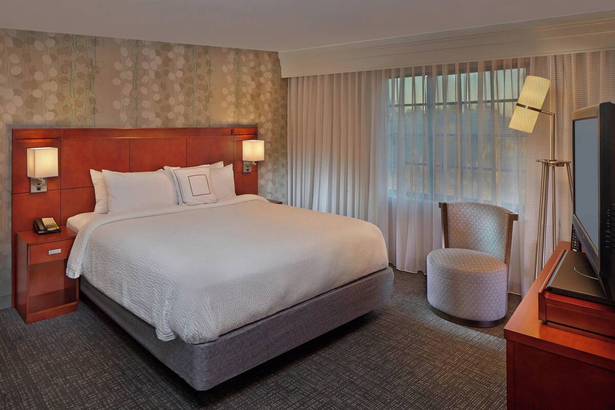 Habitación de hotel con una cama grande, dos mesitas de noche con lámparas, una silla y un televisor sobre un mueble.