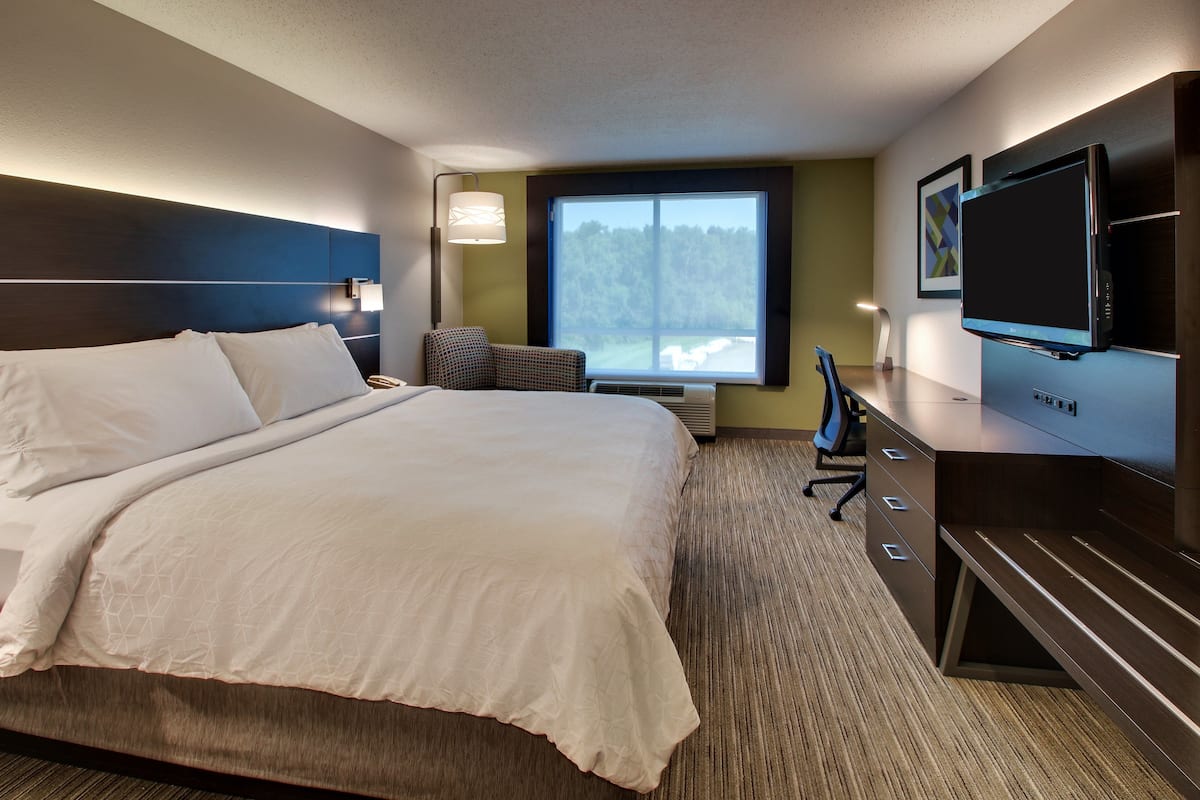 Ein Hotelzimmer mit einem großen Bett, einem Schreibtisch mit Computer, einem Fernseher und einem Fenster mit Blick auf Bäume.