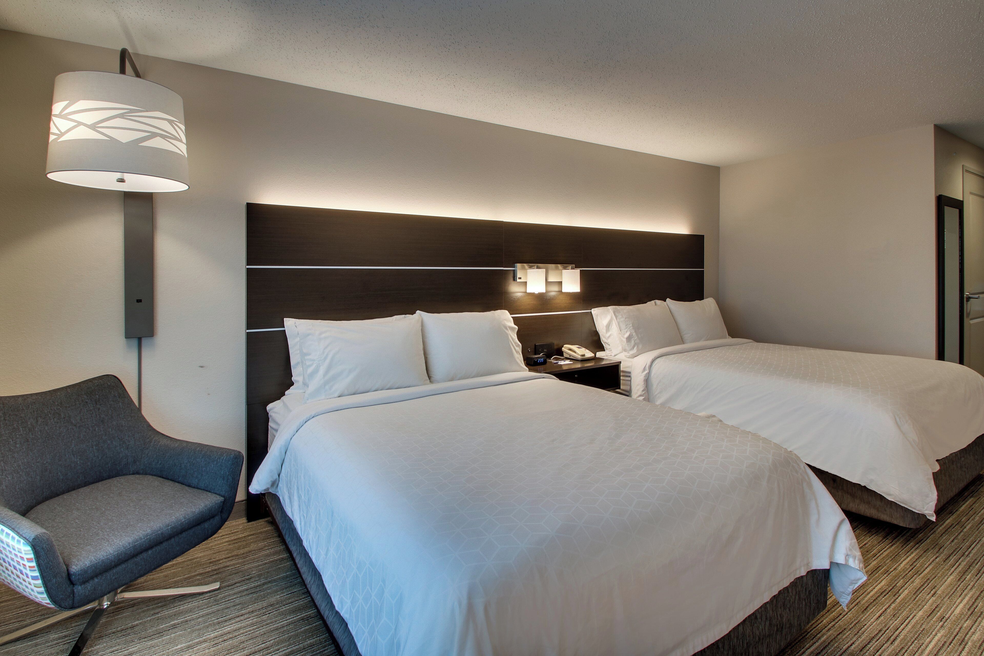 A hotel room with two beds, a chair, a nightstand with a phone, and a wall-mounted lamp.
