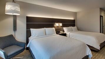 A hotel room with two beds, a chair, a nightstand with a phone, and a wall-mounted lamp.