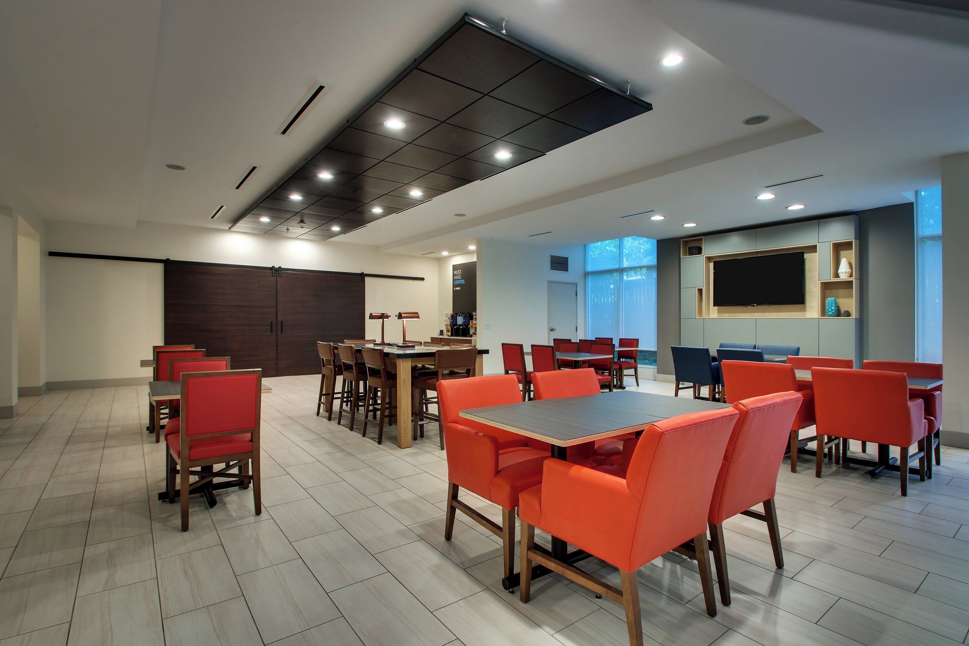 A modern dining area with multiple tables and chairs, a large flat-screen TV mounted on the wall, and a ceiling with recessed lighting.