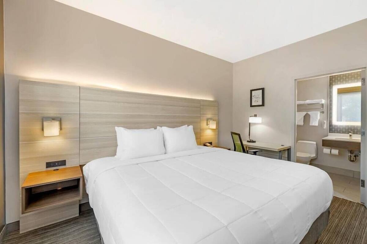 A hotel room with a large bed, bedside table, lamp, and a bathroom with a sink and mirror.