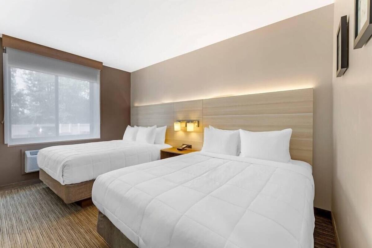 A hotel room with two beds, a large window, a wall-mounted heater, and a nightstand with a phone.