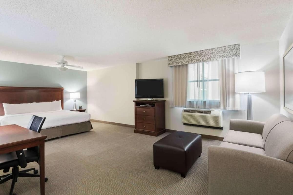 A hotel room with a bed, desk, chair, ottoman, TV, air conditioning unit, and a window with curtains.