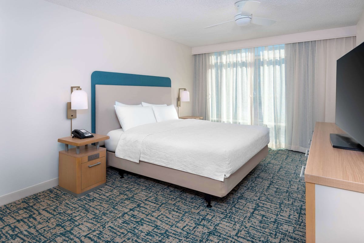 A hotel room with a bed, a desk, a chair, a nightstand, a lamp, a telephone, and a television.