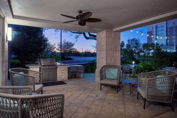 An outdoor patio area with wicker furniture, a grill, and a view of a tennis court and buildings in the background.