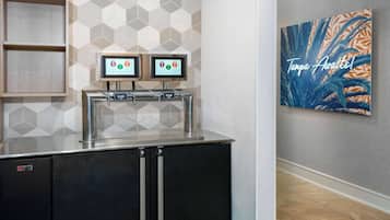 Estação de bebidas com serviço próprio, incluindo dois totens com tela sensível ao toque, um refrigerador e uma obra de arte na parede com o texto "Tampa Now!".