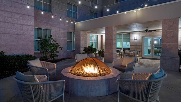 Un patio al aire libre con un pozo de fuego circular rodeado de sillas de mimbre, una zona de comedor y un edificio con grandes ventanales y paredes de piedra.