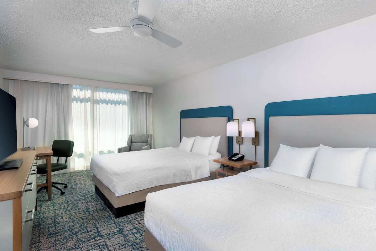 A hotel room with two beds, a desk, a chair, a desk lamp, a phone, and a ceiling fan.