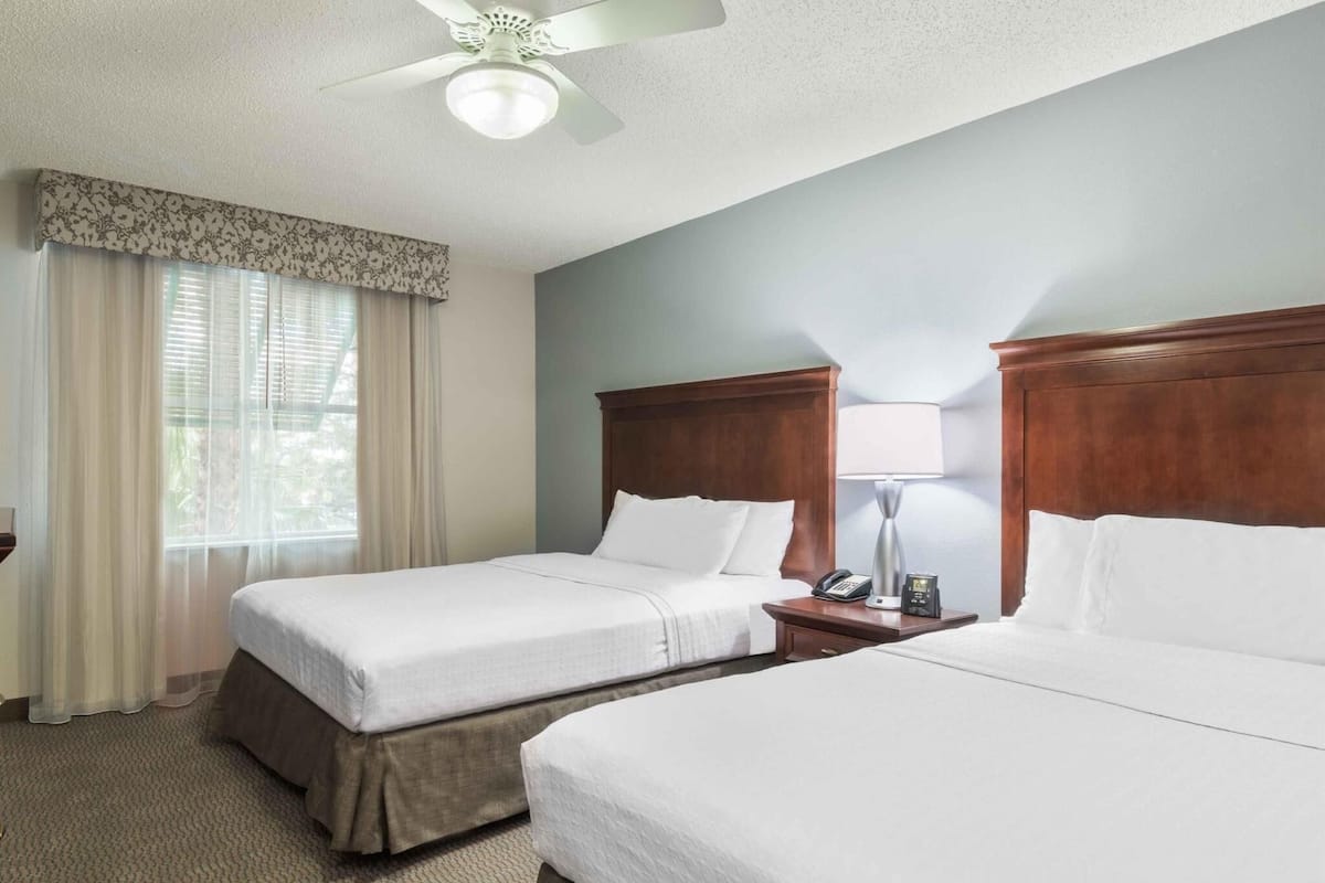 A hotel room with two beds, a ceiling fan, a nightstand with a lamp, and a window with curtains.