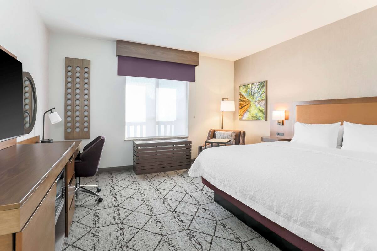 A hotel room with a large bed, a desk with a chair, a TV, a window with blinds, a lamp, and a painting on the wall.