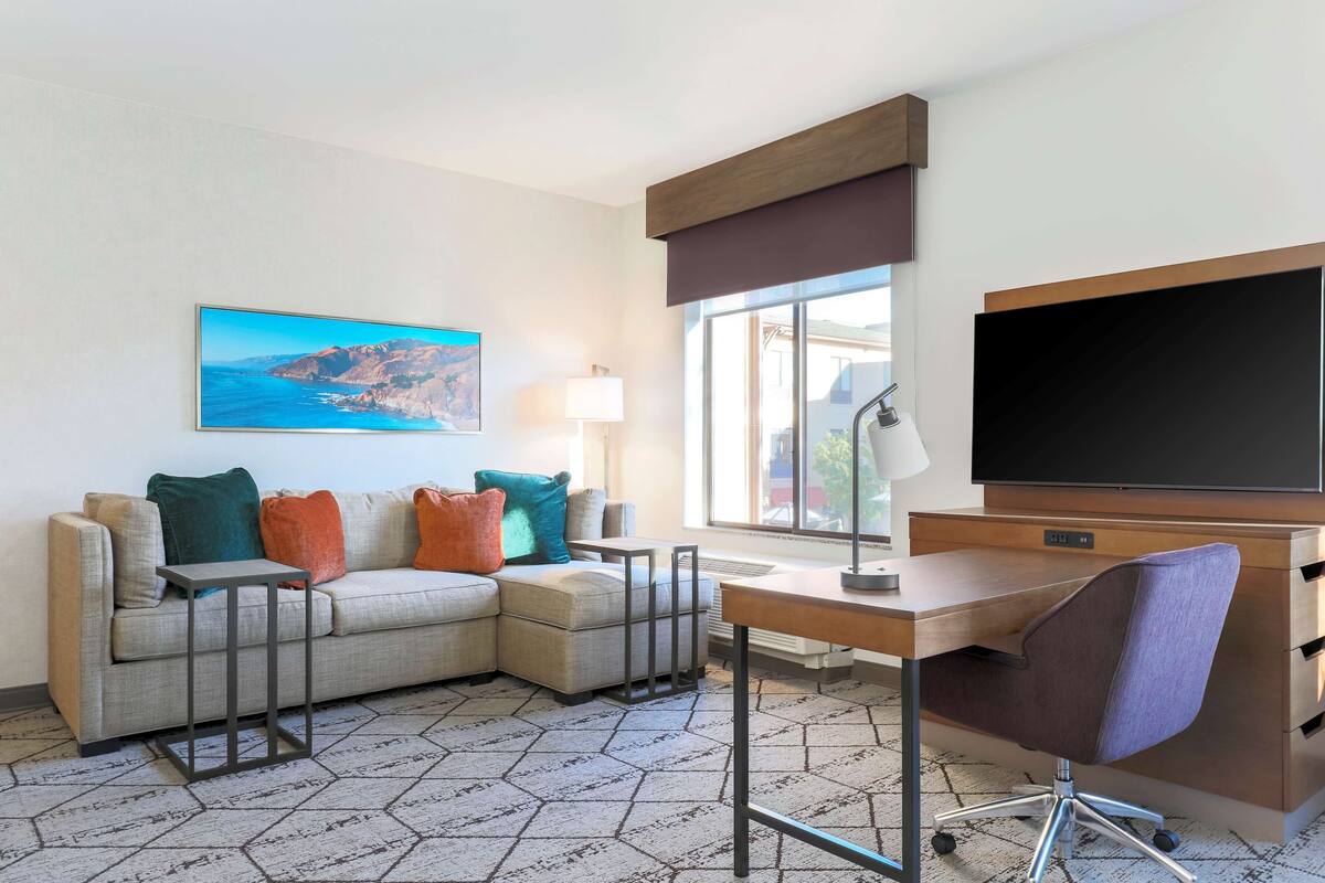 A modern hotel room with a sectional sofa, a desk with a chair, a flat-screen TV, and a large window offering a view of the outdoors.