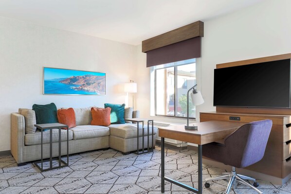 Una habitación de hotel moderna con un sofá seccional, un escritorio con silla, un televisor de pantalla plana y un ventanal con vistas al exterior.