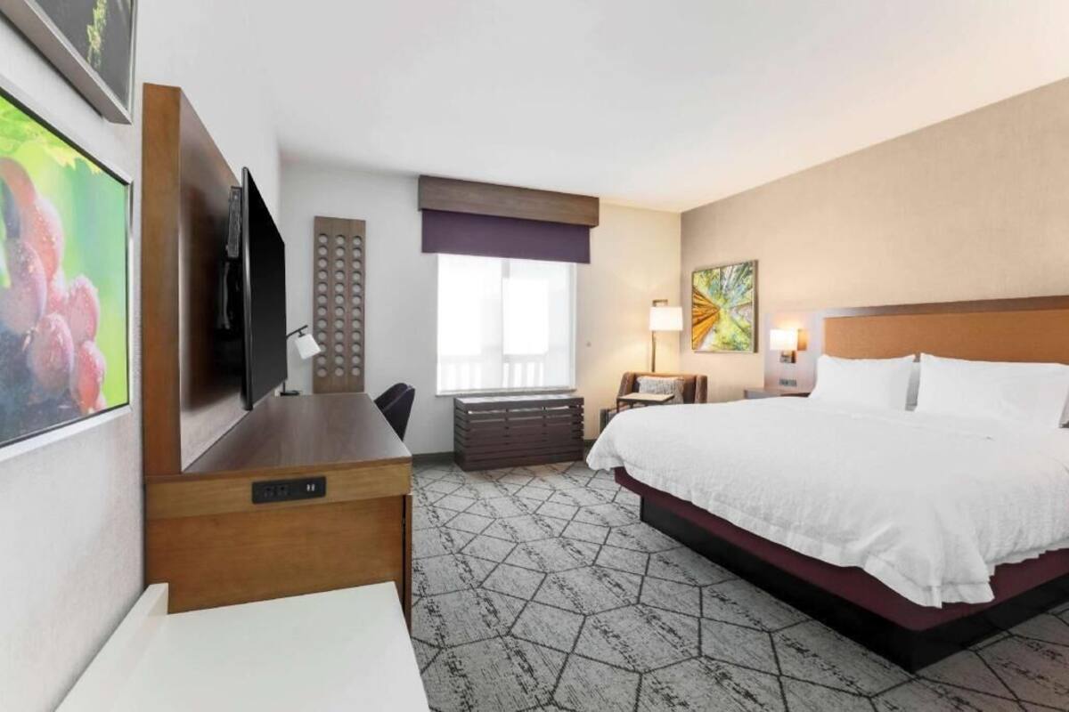 A hotel room with a bed, a desk, a television, a window, and a painting on the wall.