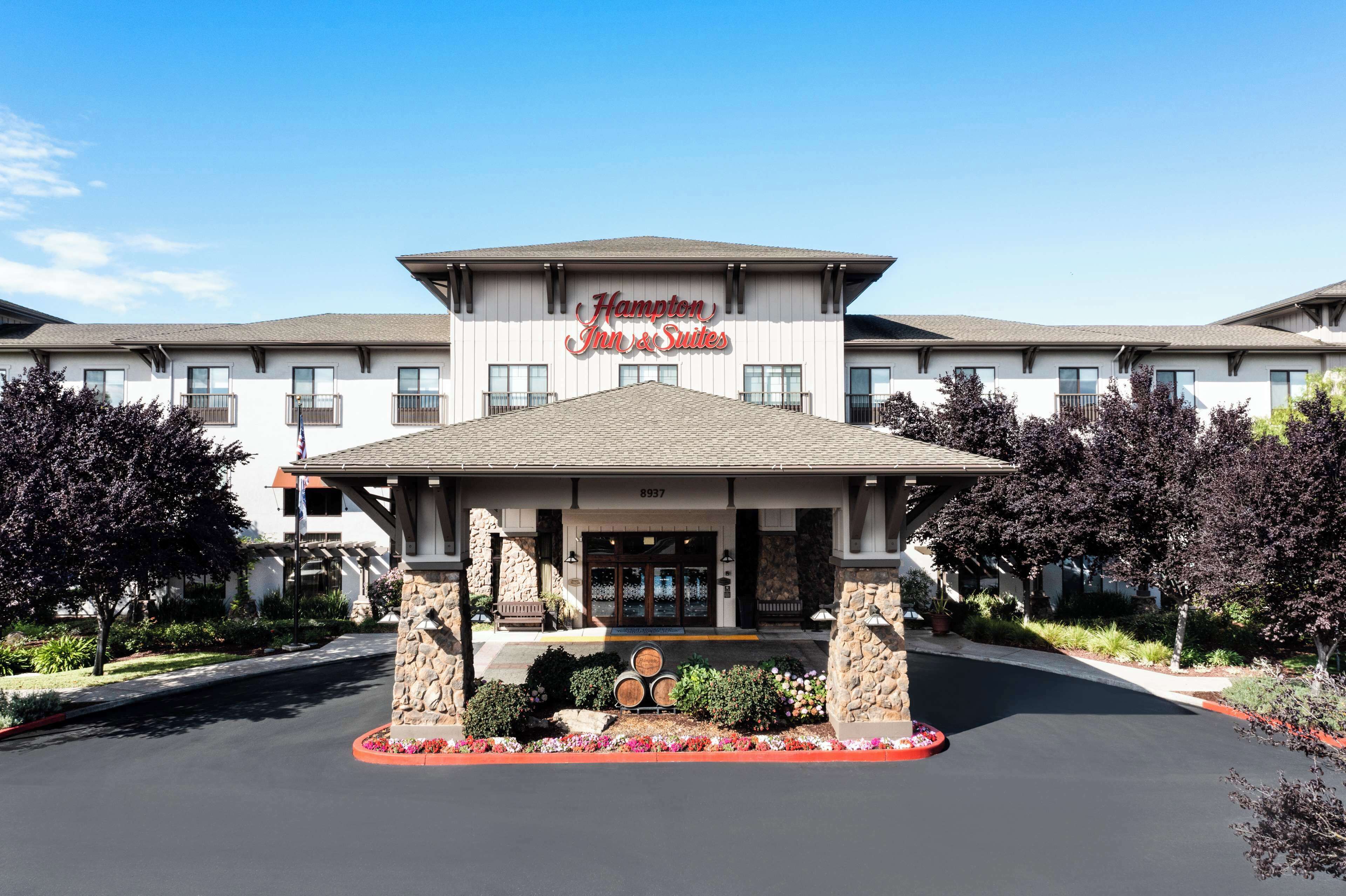 Un Hampton Inn & Suites amb façana de pedra, grans finestres i una zona d'entrada ben cuidada amb una banca i macets de flors.