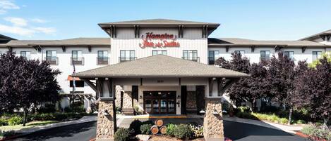 A Hampton Inn & Suites with a stone facade, large windows, and a well-maintained entrance area with a bench and flower beds.