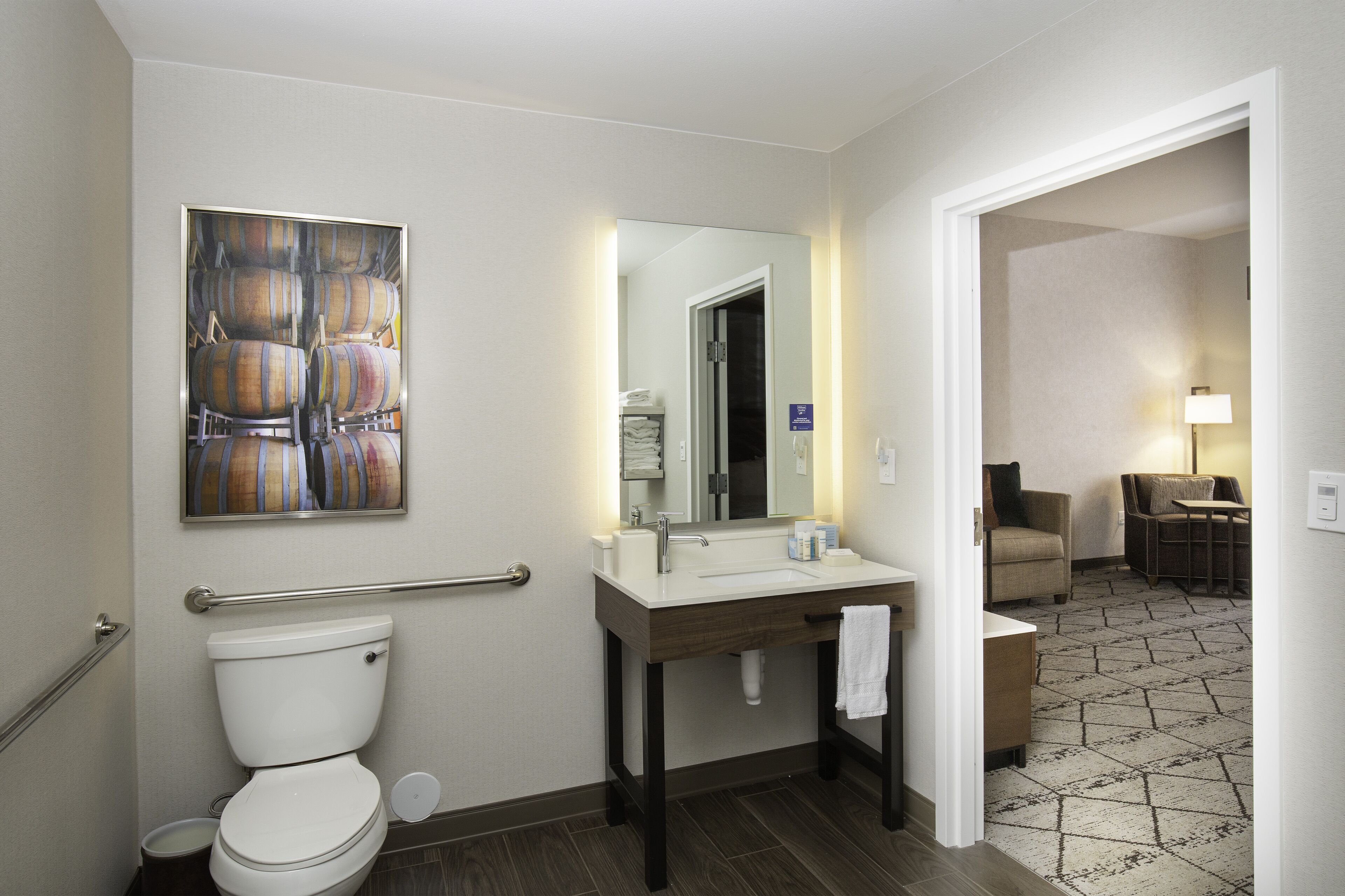 Une salle de bain moderne équipée de toilettes, d’un lavabo et d’un miroir. On y trouve une barre d’appui pour faciliter l’accès, un porte-serviettes et une petite table avec une chaise dans la pièce adjacente.