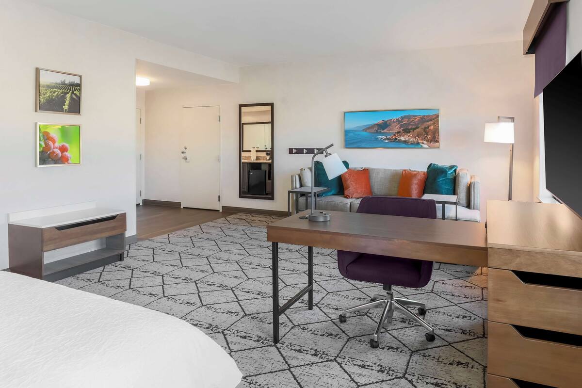 A modern hotel room with a bed, a desk, a chair, a sofa, and a large window.