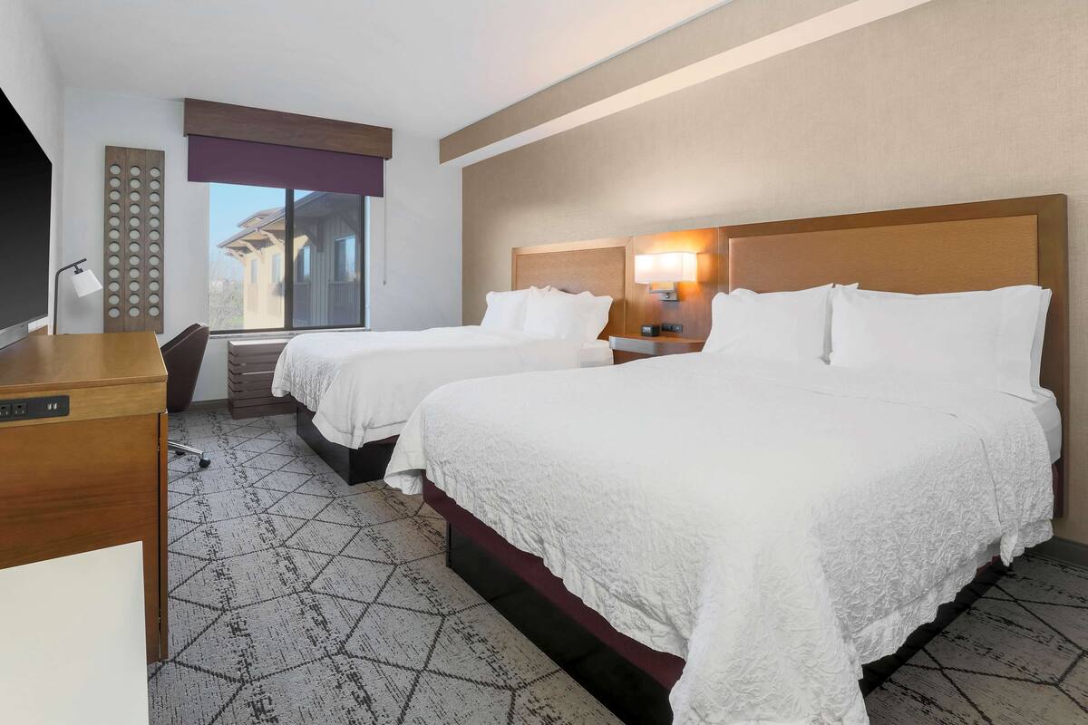 A hotel room with two beds, a desk, a chair, a lamp, and a window with a view of a building.