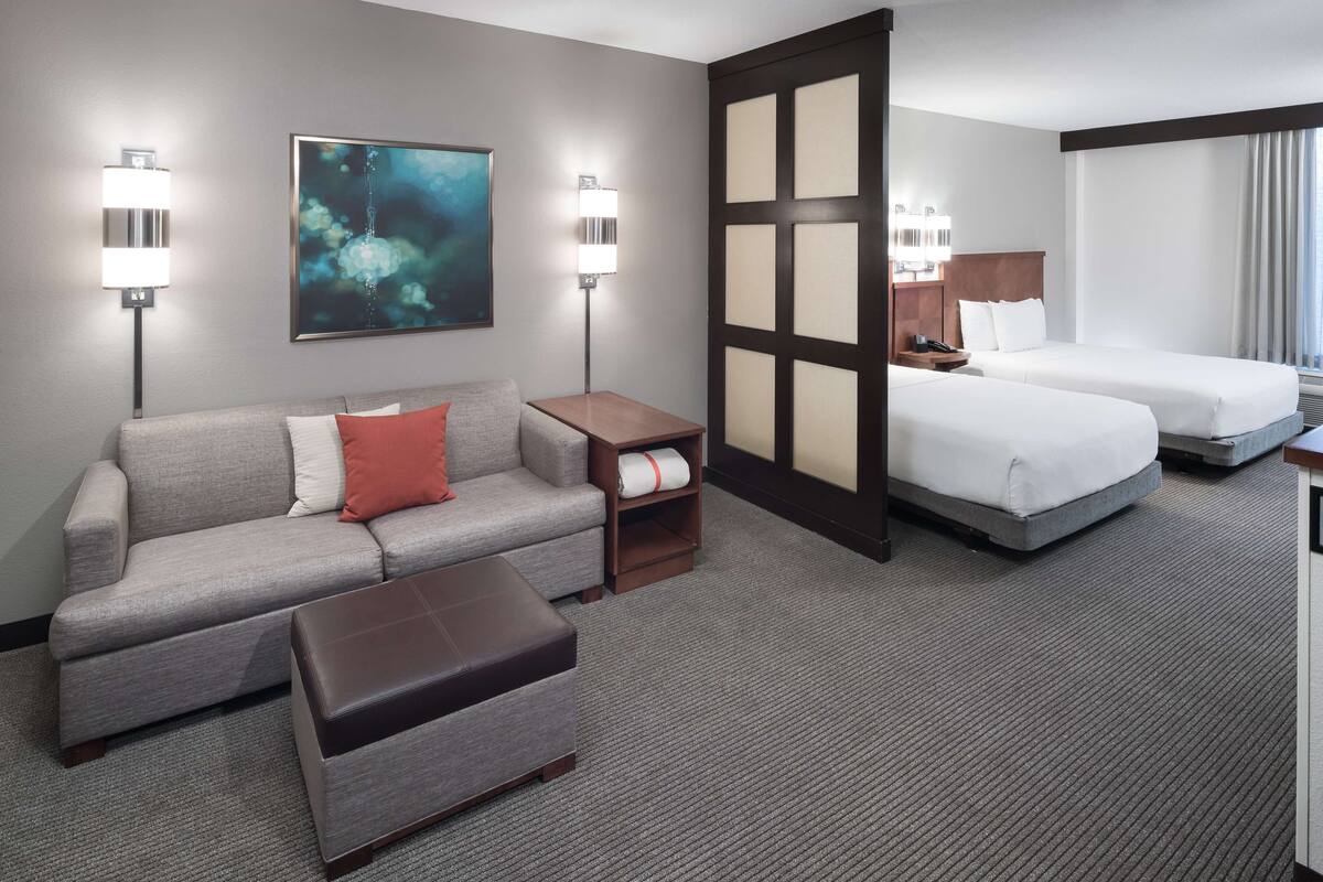 Ein Hotelzimmer mit grauem Sofa, zwei Betten, einem hölzernen Nachttisch, einem großen abstrakten Gemälde und zwei Wandleuchten.