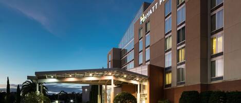 A modern hotel entrance with a covered walkway, a parking lot, and a clear sky.
