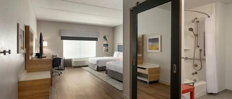 A modern hotel room with a wooden floor, a bed, a desk, a chair, a wardrobe, a bathroom with a shower, and a large mirror.