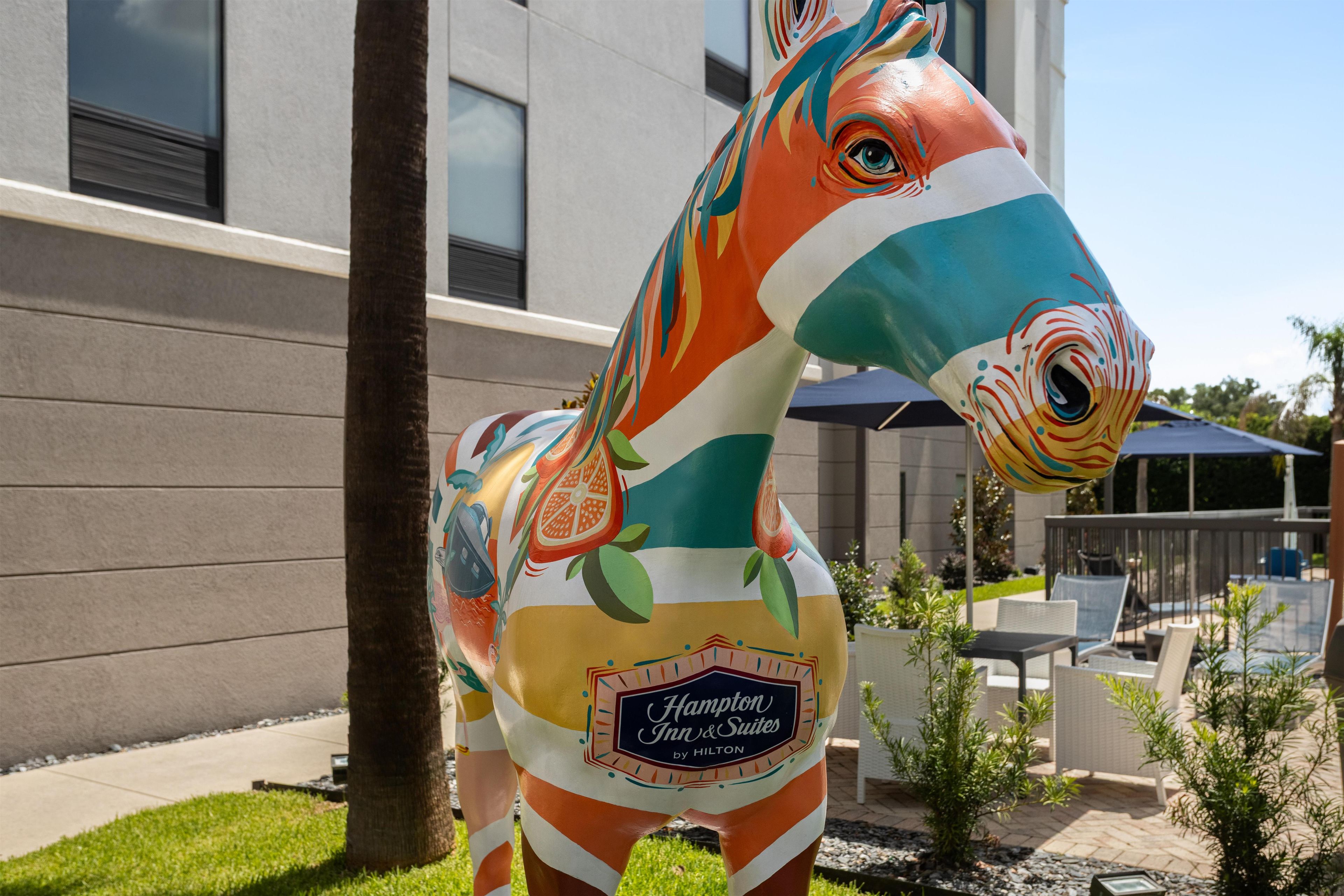 A colorful horse statue with a Hampton Inn & Suites logo, located outdoors with a building and patio furniture in the background.