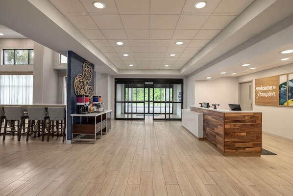 Zona de recepción con un mostrador de madera, un área de estar con taburetes y una máquina de café instalada en la pared.