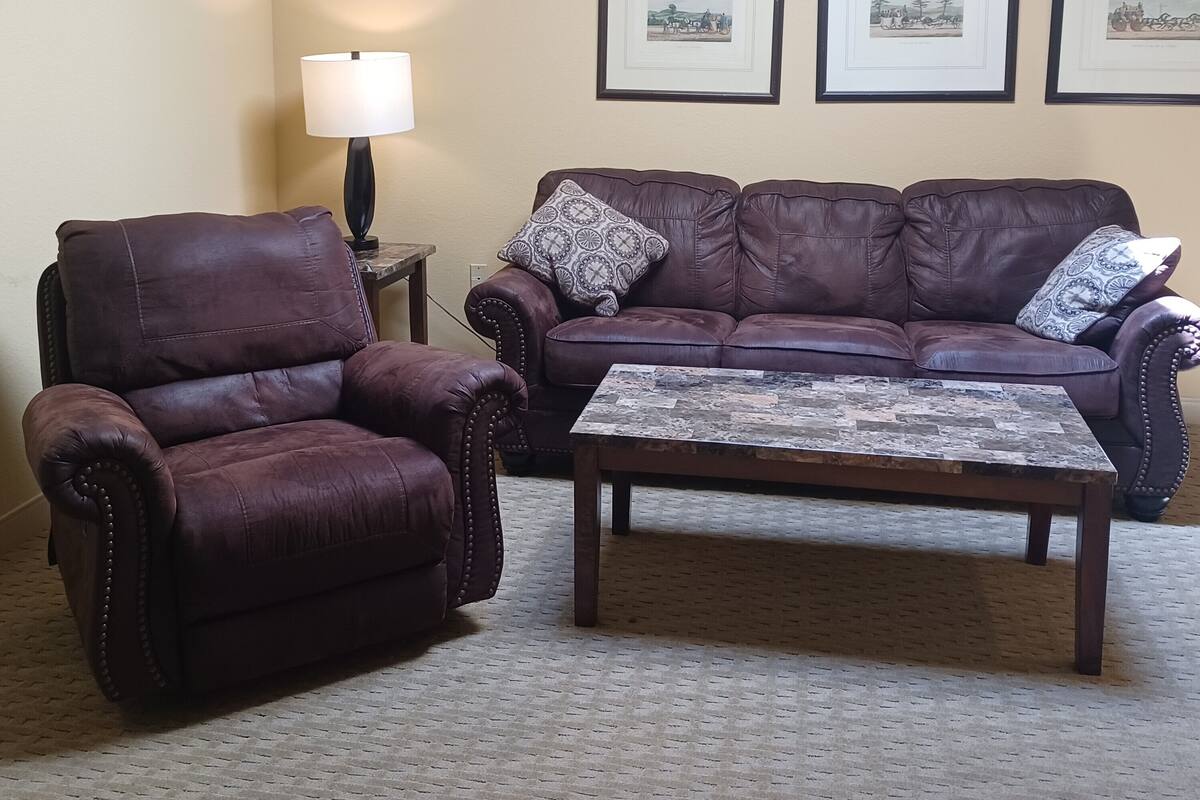 A living room with a brown leather armchair, a brown leather sofa, and a coffee table. There are three framed pictures on the wall.
