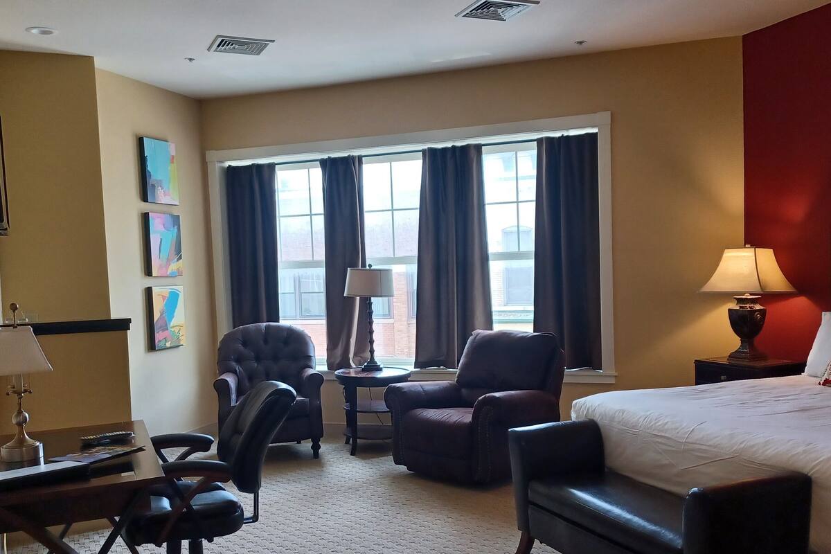 A hotel room with a bed, two armchairs, a desk with a lamp, a window with curtains, and artwork on the wall.