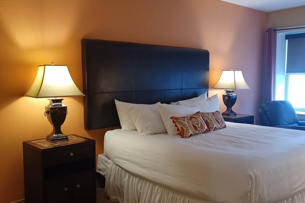 A hotel room with a large bed, two bedside tables with lamps, a dark headboard, and a window with curtains.