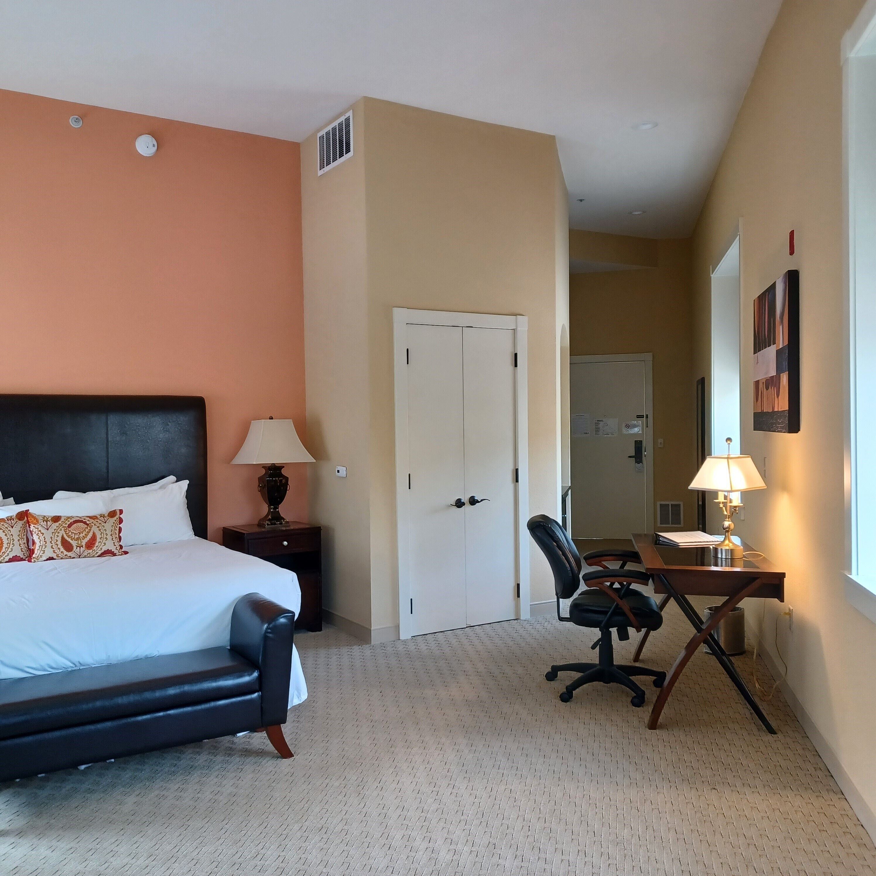 A hotel room with a bed, a desk with a chair, a lamp, a door, and a window.