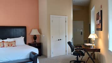 A hotel room with a bed, a desk with a chair, a lamp, a door, and a window.