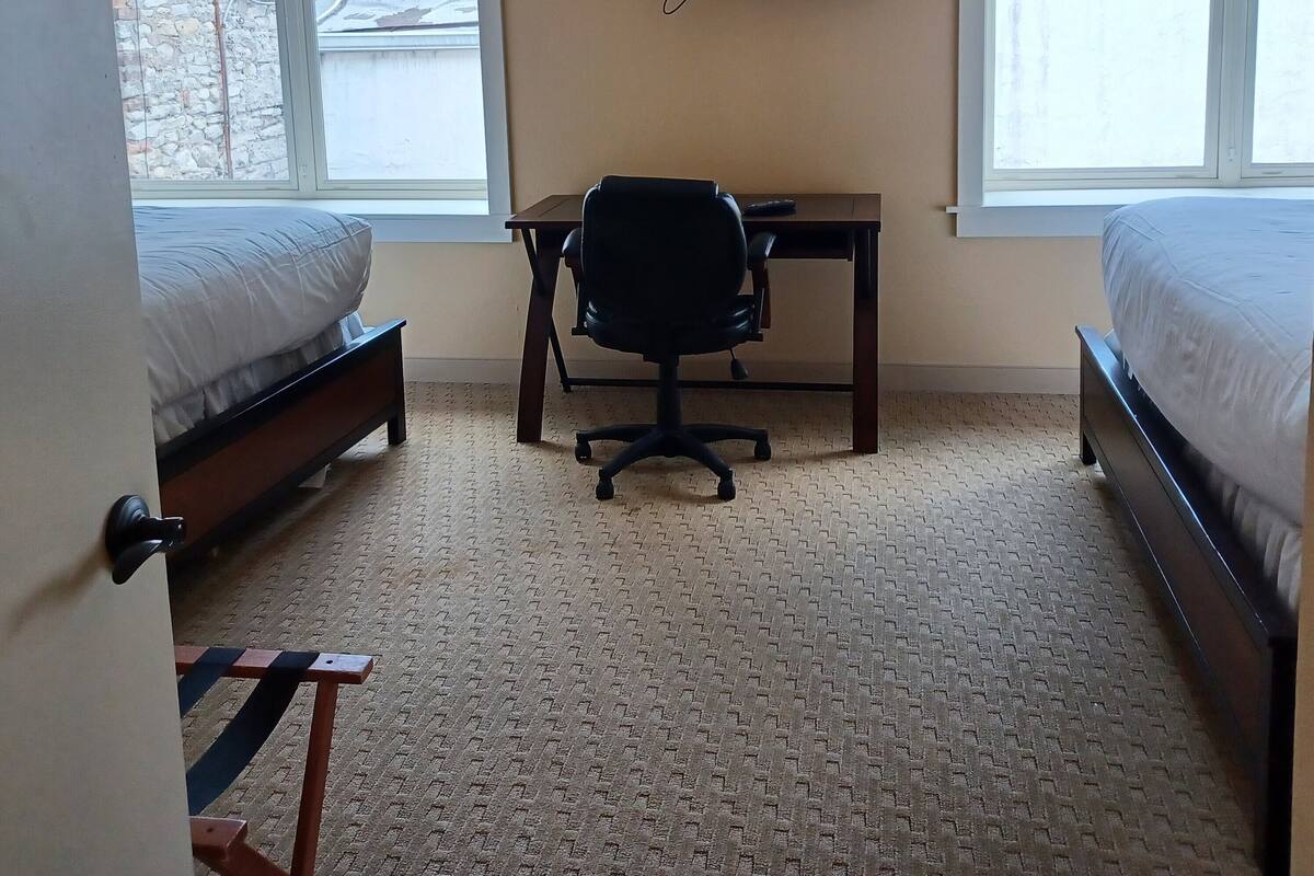 A hotel room with two beds, a desk, a chair, a TV, and large windows.