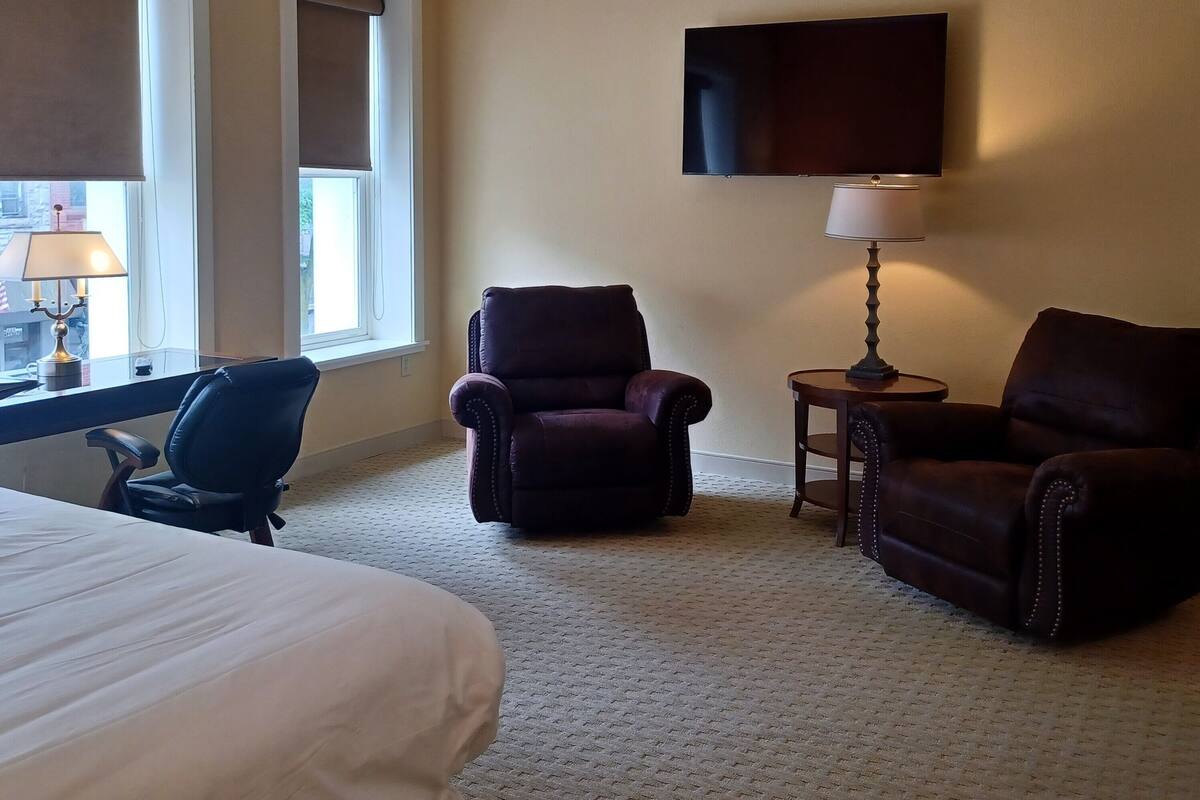 A hotel room with a bed, two armchairs, a small table with a lamp, a flat-screen TV mounted on the wall, and a window with blinds.