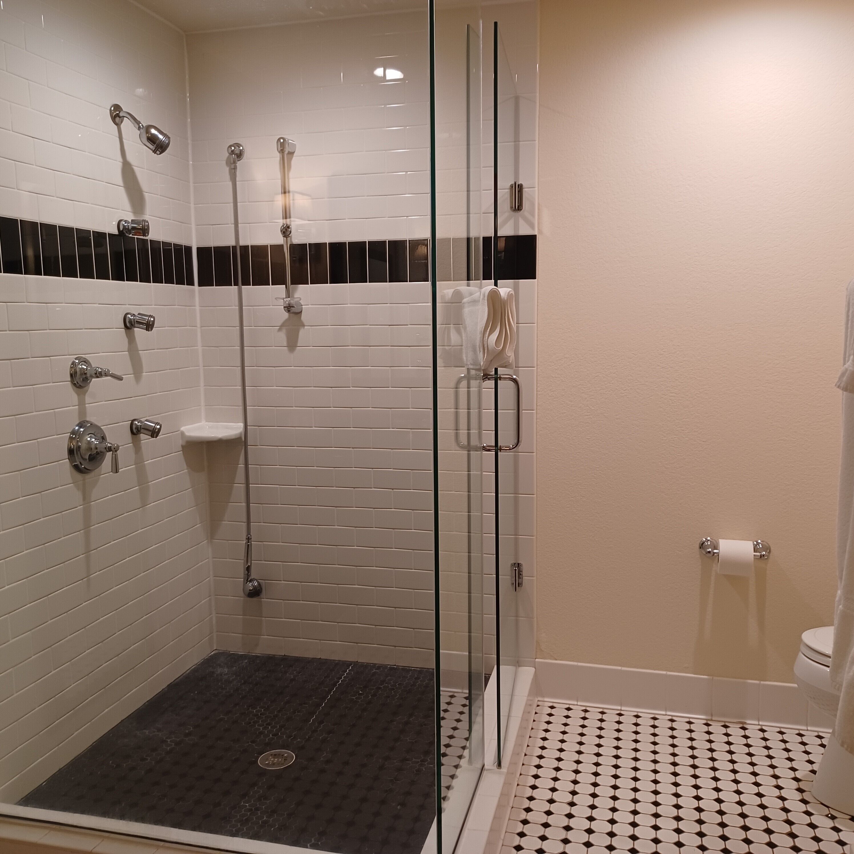 Et moderne badeværelse med en brusekabine med glasvægge, hvid klinkegulv, et gulv med et sort-hvidt mønster og et toilet med en håndklædeholder.