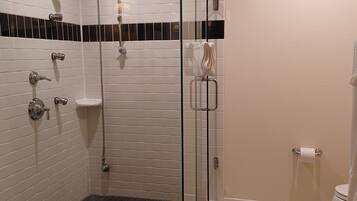 Un baño moderno con ducha envuelta en cristal, paredes con azulejos blancos, piso con estampado blanco y negro, y un inodoro con dispensador de toallas de papel.