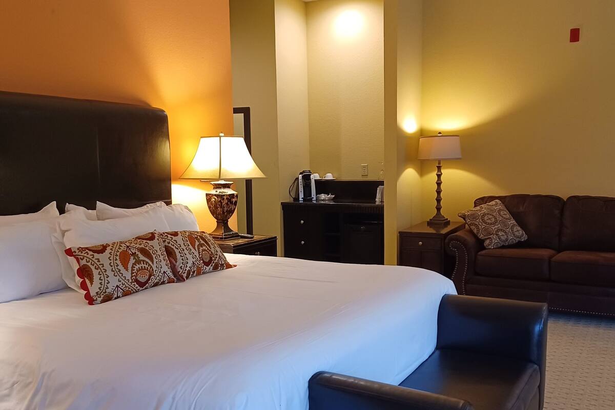 A hotel room with a large bed, two bedside tables with lamps, a dark-colored sofa, and a small table with a coffee maker.