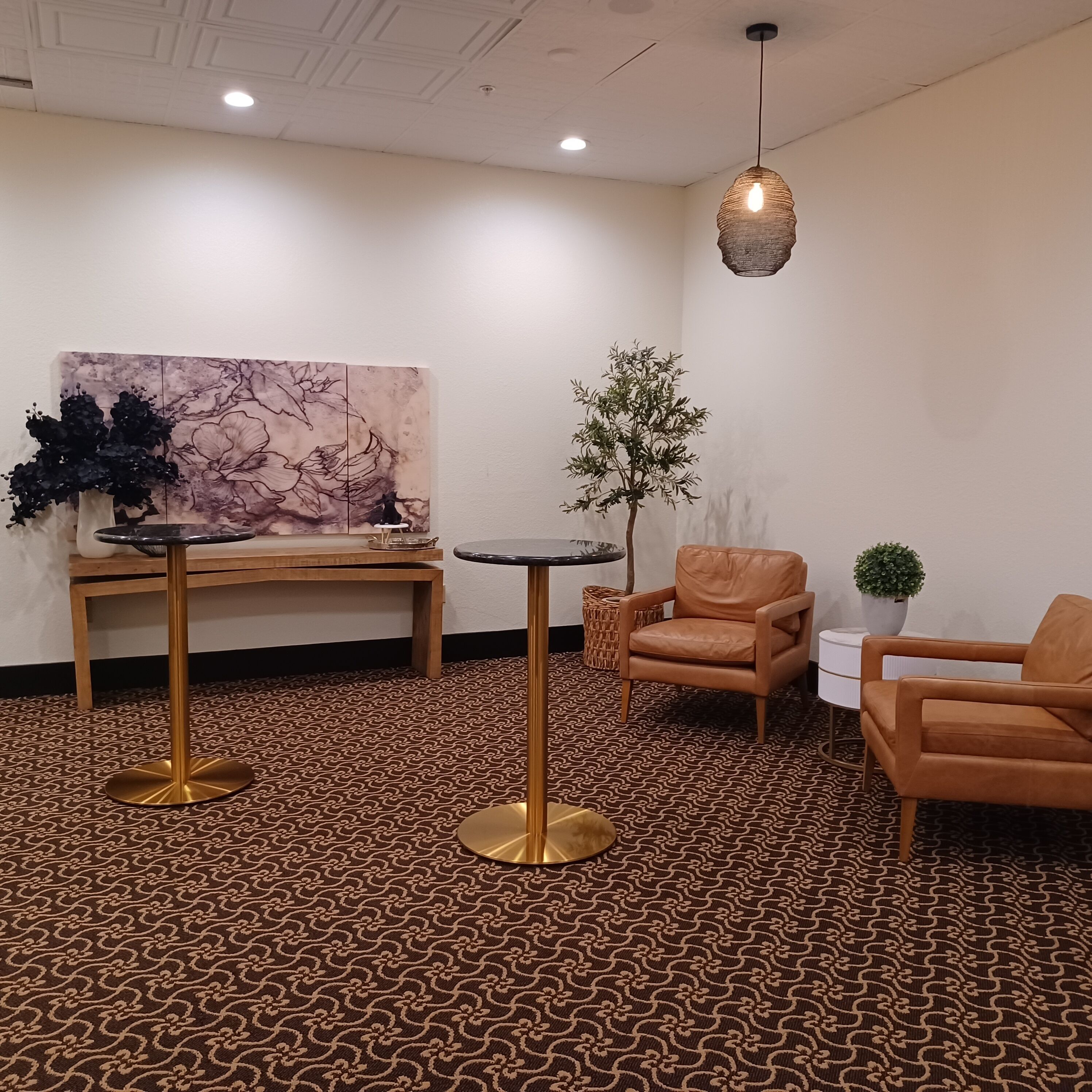 A room with a patterned carpet, a painting on the wall, a table with a vase, a chair, and a hanging light fixture.