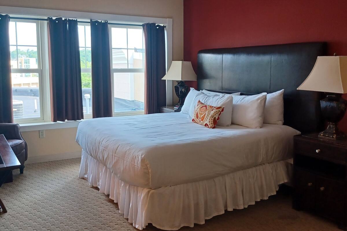 A hotel room with a large bed, two bedside lamps, a window with curtains, a chair, and a view of buildings outside.