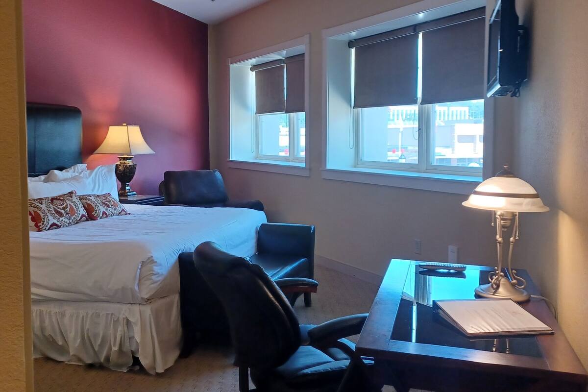 A hotel room with a bed, desk, chair, lamp, and windows.