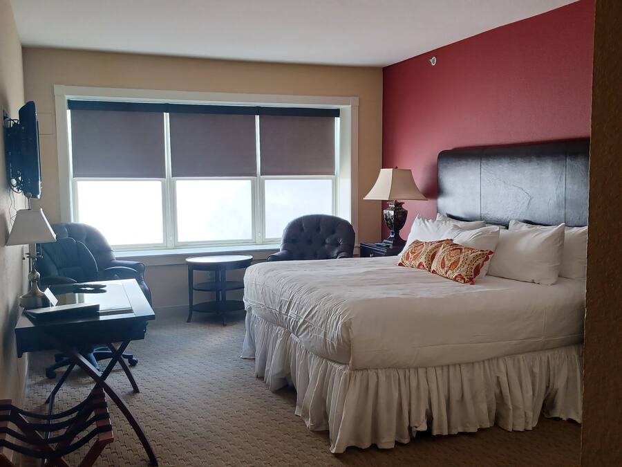 A hotel room with a large bed, a desk with a chair, a small round table, and a window with blinds.