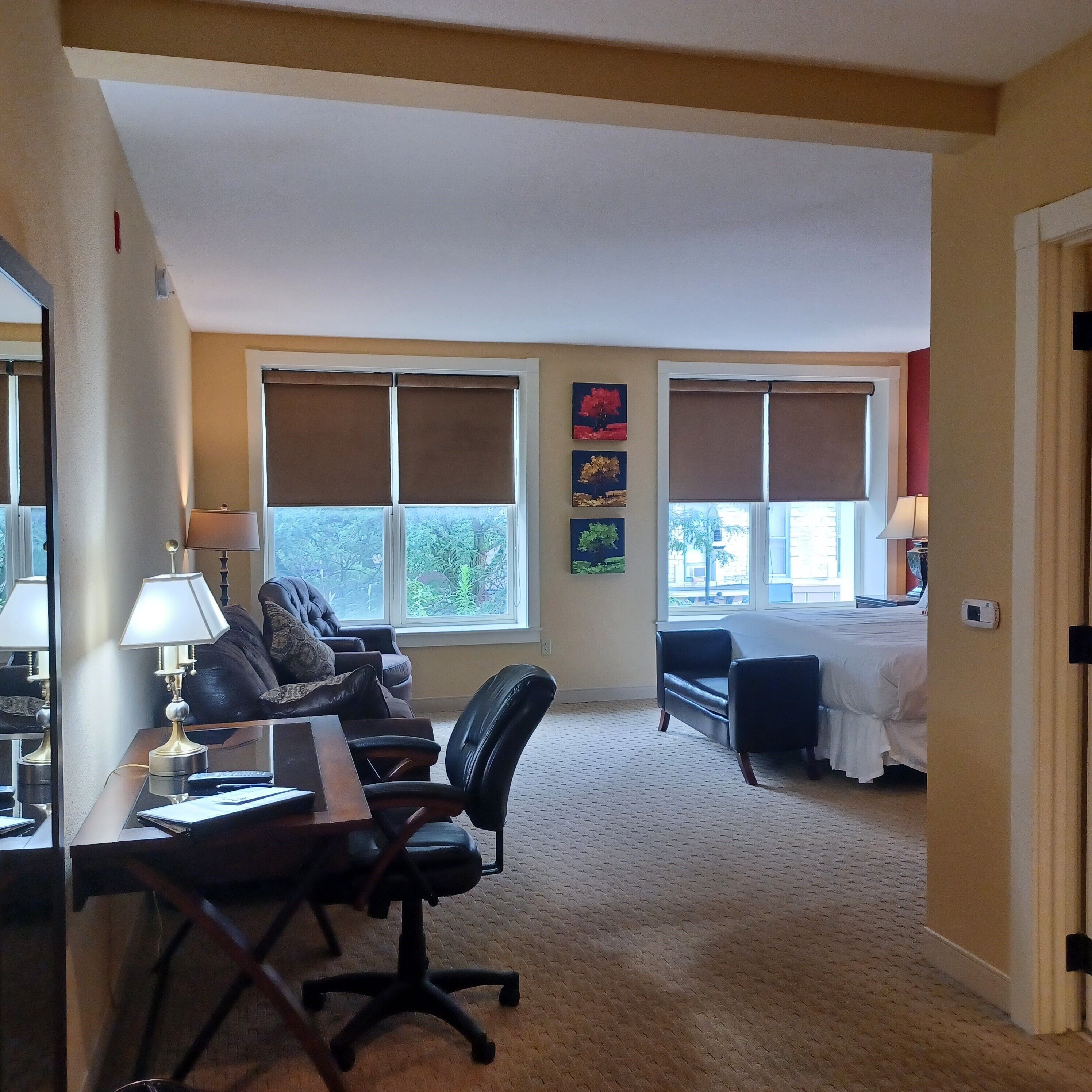 A hotel room with a desk, office chair, two windows with blinds, a bed, and a small couch.