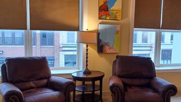 A room with two leather armchairs, a small round table, and a lamp. There are large windows with blinds, and two pieces of abstract art on the wall.