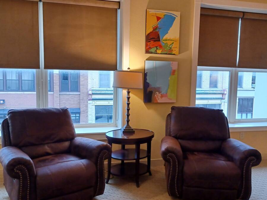 A room with two leather armchairs, a small round table, and a lamp. There are large windows with blinds, and two pieces of abstract art on the wall.