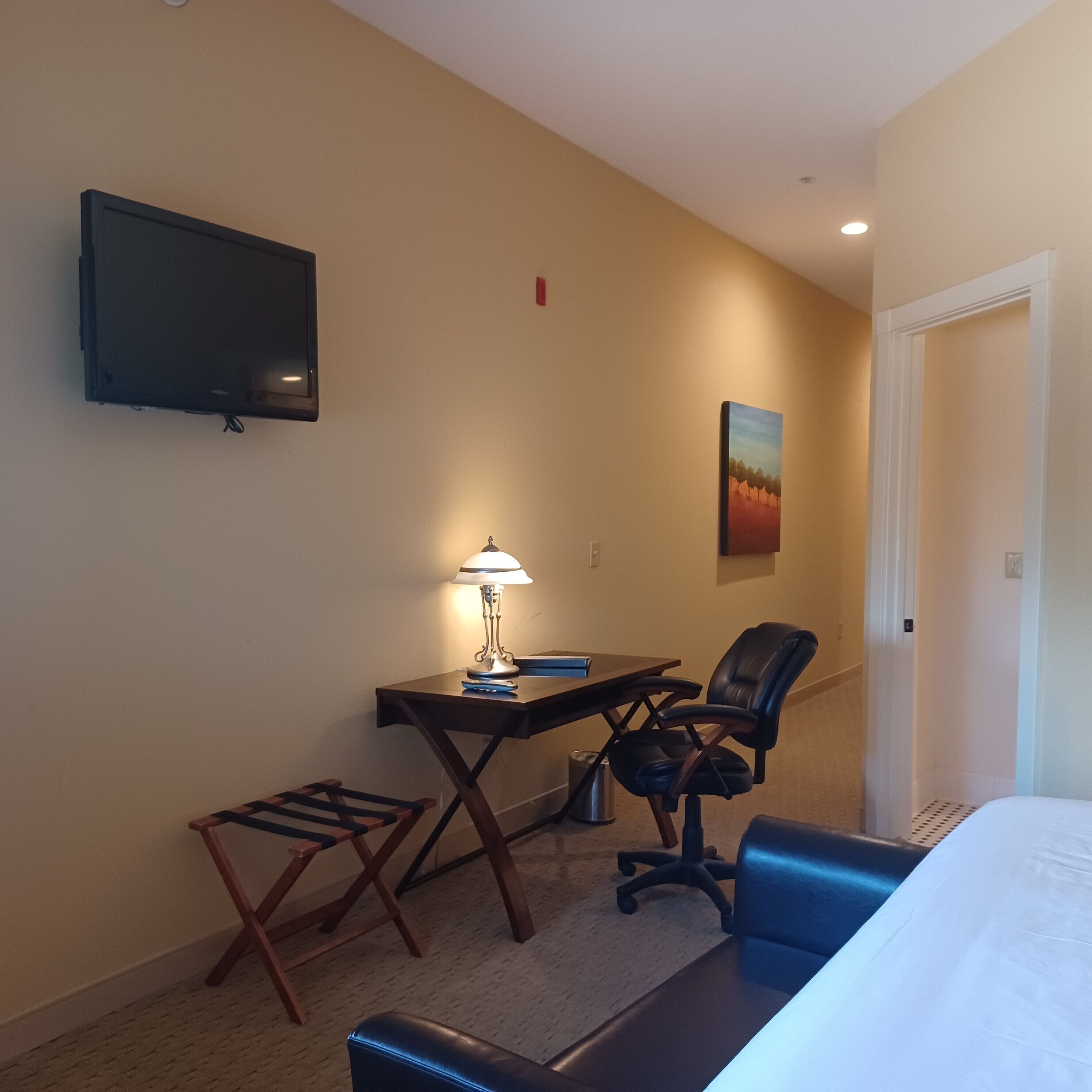 A hotel room with a bed, a desk with a chair, a TV mounted on the wall, and a painting on the wall.