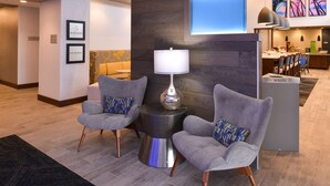 Un vestíbulo con dos sillones grises, una mesita metálica con lámpara y un mostrador de recepción con pantalla digital.